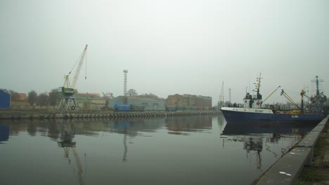 Barco-De-Pescador-Azul-Atracado-En-El-Puerto-De-Liepaja-En-Un-Día-De-Niebla,-Vagones-De-Carga-Seca-Y-Grúa-Portuaria-En-El-Fondo,-Reflejos-En-El-Agua,-Tiro-Ancho