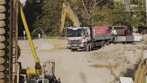 Excavator-unloading-dirt-into-a-truck-on-a-construction-site-in-Vienna,-Austria-in-slow-motion-with-a-worker-passing-by