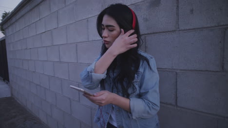 Mujer-Joven-Mirando-Su-Teléfono-En-El-Callejón,-Junto-A-La-Pared,-Con-Auriculares-Rojos---Plano-Medio