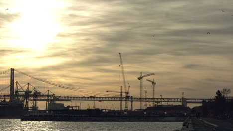 Puente-Del-25-De-Abril-Al-Atardecer-Con-Tráfico-Rodado,-Grúas-De-Construcción-Y-El-Río-Tajo