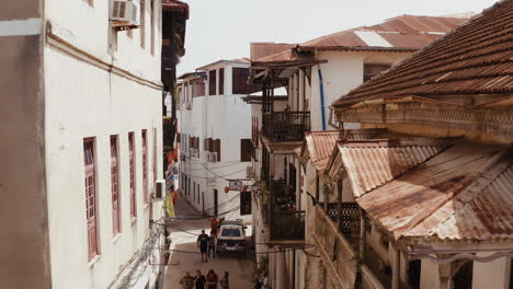 Antigua-Calle-Estrecha-De-La-Ciudad-De-Piedra-De-Zanzíbar-Con-Coches-Aparcados-A-Los-Lados-De-La-Carretera-Y-Turistas-Y-Locales-Caminando-Por-La-Calle