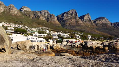 Toma-Estática-De-Casas-De-Vacaciones-En-La-Base-De-La-Montañosa-Y-Rocosa-Cordillera-De-Los-Apóstoles-Sudafricanos-Con-Paisaje-Marino-Debajo