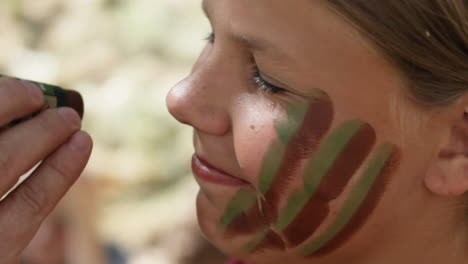 Un-Primerísimo-Plano-De-Una-Chica-Sonriente-Con-La-Cara-Pintada-Con-Pintura-De-Guerra-De-Colores-Verde-Y-Marrón-Por-Otra-Persona