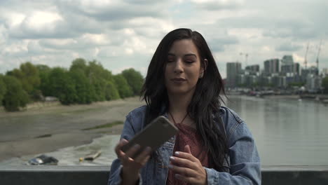 Brunette-Latina-tourist-taking-a-selfie,-posing-doing-victory-sign-with-her-hand,-while-standing-on-the-railing-of-a-bridge-in-London