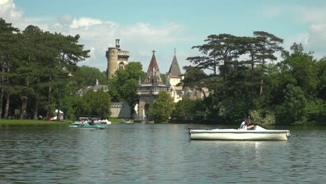 Blick-Auf-Das-Schloss-Laxenburg-Hinter-Dem-See-Mit-Vorbeifahrenden-Booten-Im-Vordergrund-An-Einem-Hellen-Und-Sonnigen-Sommertag