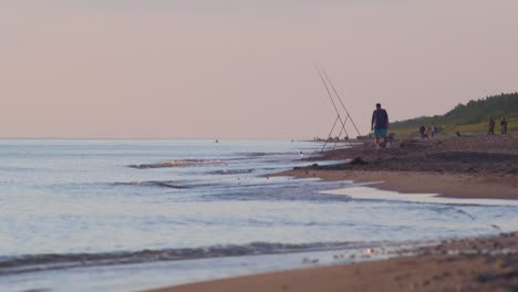 Un-Pescador-Pesca-Peces-Planos-Con-Una-Caña-De-Pescar-En-La-Costa-Del-Mar-Báltico-En-La-Soleada-Tarde-De-Verano-Antes-Del-Atardecer,-Tiro-Amplio-Desde-La-Distancia