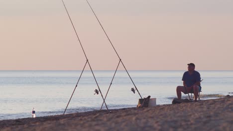 Un-Pescador-Pesca-Peces-Planos-Con-Una-Caña-De-Pescar-En-La-Costa-Del-Mar-Báltico-En-La-Soleada-Tarde-De-Verano-Antes-Del-Atardecer,-Tiro-Medio-Desde-La-Distancia