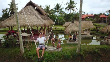 Drone-Shot-of-a-tourist-enjoying-a-swing-between-two-coconut-trees-that-swings-out-over-some-Rice-Terraces-in-Bali,-Indonesia