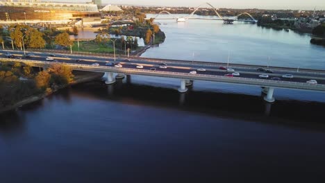 Aerial-shot-pulling-back-from-a-bridge-to-reveal-the-landscape-and-stadium-view-from-above,-with-traffic-during-golden-hour