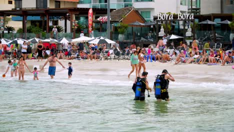 Two-male-divers-walking-towards-the-shore-and-people-resting-in-Bombas-and-Bombinhas-beaches,-Brazil