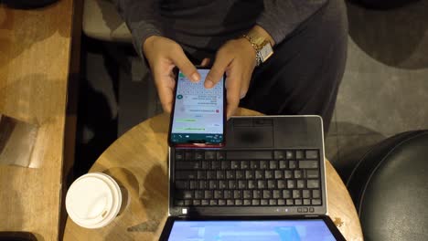 Top-view-of-business-woman-hands-texting-on-smartphone-with-laptop-and-coffee-on-the-table-and-yellow-light-ambiance-from-coffee-shop