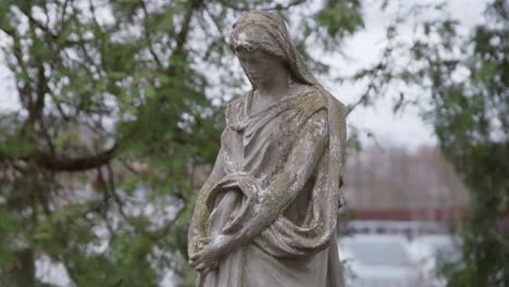 Vieja-Escultura-De-Aspecto-Triste-De-Piedra-De-La-Madre-María-En-El-Antiguo-Cementerio-Con-árboles-Ondeando-En-El-Viento