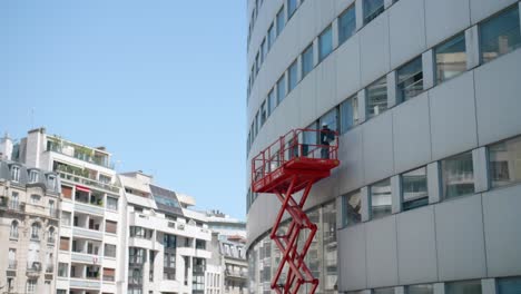 Trabajador-En-Plataforma-Elevada-Trabajando-En-La-Ventana-En-El-Exterior-Del-Edificio-Maison-De-La-Radio-En-París,-Francia