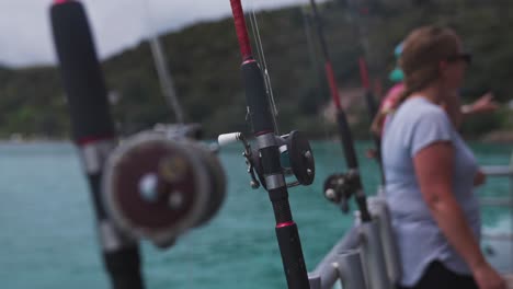 Fishing-rods-lined-up-on-side-of-boat-with-out-of-focus-people-relaxing,-recreation-trip