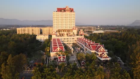 Hotel-Resort-and-Spa-Exterior-Orbit-Pan-Drone-Shot-During-Sunrise-with-Mountainous-Background-Surrounded-by-Trees-Along-the-Beach-of-Cha-Am-in-Thailand