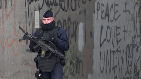 Un-Oficial-De-Policía-De-Crs-Armado-Con-Un-Rifle-De-Asalto-Hace-Guardia-Frente-A-Un-Grafiti-En-Un-Puente-Que-Dice:-“paz,-Amor,-Unidad”