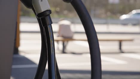 Electric-car-cables-against-person-sitting-on-station-bench-using-smartphone-while-waiting-for-charging-his-vehicle