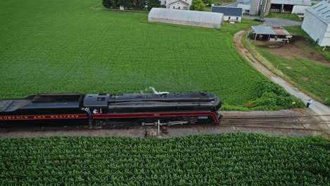 Una-Vista-Aérea-Paralela-De-Un-Tren-De-Pasajeros-De-Vapor-Antiguo-Que-Sopla-Humo-A-Través-De-Tierras-Agrícolas-Amish