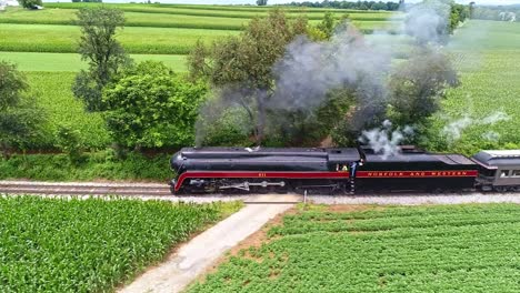 Una-Vista-Aérea-Paralela-De-Un-Tren-De-Pasajeros-De-Vapor-Antiguo-Que-Viaja-Con-Humo-Negro-A-Través-De-Tierras-De-Cultivo