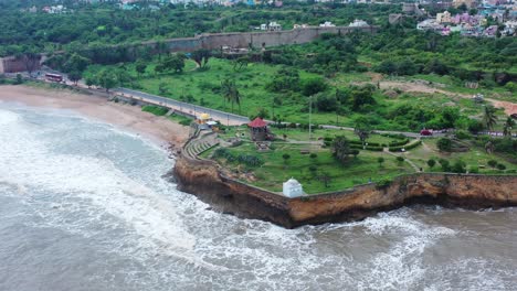 footage-shot-from-over-the-ocean-zooming-into-a-cliffside-structure,-foamy-white-waves-gently-lapping-the-shore,-cliffside-rocky-side,-greenery,-people-walking-around,-top-shot-of-structure,-buildings