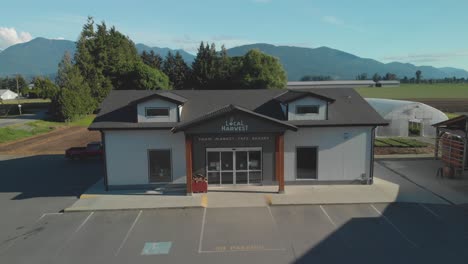 Local-Harvest-Market-and-farm-store-front-aerial-from-parking-lot-ascending-while-panning-down-tracking-the-building-within-frame-bright-sunny-spring-day-blue-sky-trees-mountains-in-background