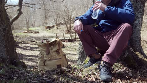 Älterer-Mann-Mit-Rucksack-Sitzt-Am-Baum-Im-Wald-Und-Trinkt-Wasser