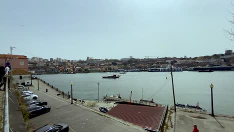 Un-Barco-Navegando-Por-Las-Plácidas-Aguas-De-Oporto,-Portugal,-Con-Vistas-A-Los-Coches-Aparcados-Y-A-La-Gente-En-Primer-Plano-Y-Las-Estructuras-De-La-Ciudad-Costera-Al-Fondo