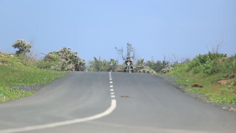 on-ground-slowed-view-of-man-in-dark-clothes-on-a-bike-approaching-towards-the-camera-from-the-other-direction,-bright-blue-clear-sky-in-background,-grass-and-trees-on-both-sides-of-road,-grey-roads