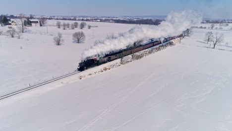 Vista-Aérea-De-Una-Antigua-Locomotora-De-Vapor-Que-Se-Acerca-Tirando-De-Automóviles-De-Pasajeros-Y-Soplando-Humo-Y-Vapor-Después-De-Una-Tormenta-De-Nieve