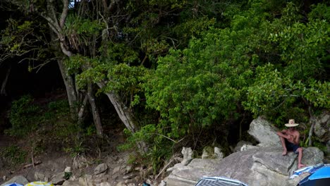 Jib-up-of-families-relaxing-in-the-sand-under-umbrellas-on-a-rocky-shore-near-woods-in-Praia-da-Sepultura,-Brazil