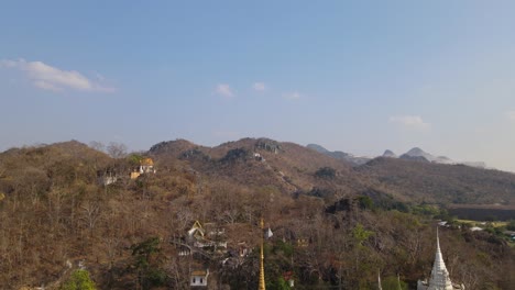 Wat-Phra-Phutthabat,-Saraburi,-Thailand,-descending-aerial-shot-revealing-limestone-mountains,-brown-trees-scorched-by-the-summer-sun-and-temple-stairway,-the-golden-Buddhist-temple,-and-its-complex