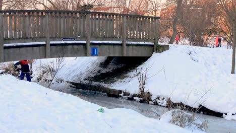 Kleiner-Junge,-Der-Auf-Schlittschuhen-Unter-Einer-Brücke-In-Einer-Schneelandschaft-Vorbeifährt,-Um-Auf-Die-Andere-Seite-Zu-Gelangen,-Mit-Einem-Kleinen-Schild,-Das-Den-Verkehr-Regelt,-Wer-Bei-Gleichzeitiger-Ankunft-Zuerst-Fährt