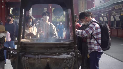 Menschen-Mit-Gesichtsmaske,-Während-Sie-Sich-Im-Senso-ji-In-Japan-Mit-Rauch-Reinigen