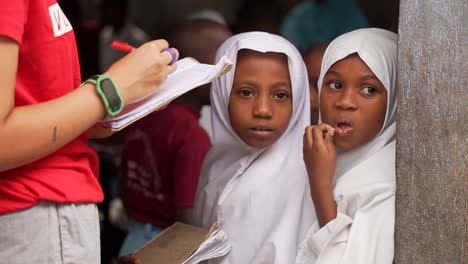 Weiße-Kaukasische-Ehrenamtliche-Lehrerin-Korrigiert-Die-Hausaufgaben-Afrikanischer-Mädchen-Mit-Weißem-Kopftuch,-Die-In-Der-Schule-Lernen