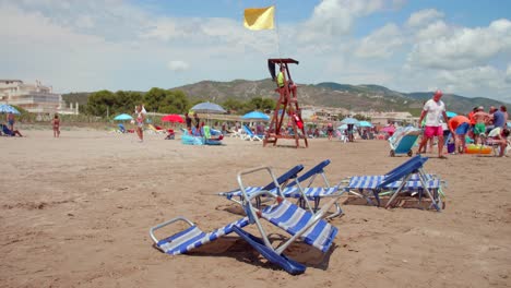 Sillas-De-Playa-Vacías-En-La-Arena-Con-Bandera-Amarilla-Ondeando-En-El-Puesto-De-Socorrista-En-Alcossebre,-Costa-Del-Azahar,-España