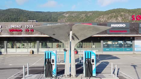 Cargador-Artístico-De-Alta-Velocidad-Para-Coches-Eléctricos---Estación-De-Carga-Mer-En-Flaa-Noruega---Antena-Que-Se-Mueve-Lentamente-Hacia-Arriba-Con-Un-Centro-Comercial-En-Segundo-Plano