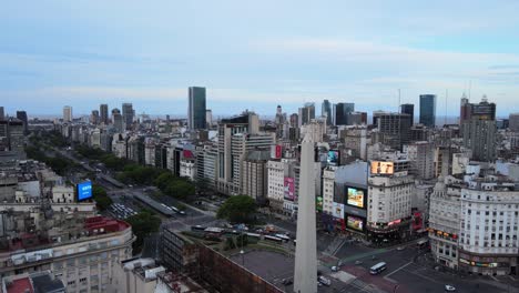 Cinematic-low-level-aerial-pull-out-shot-slowly-flying-over-a-chalet-and-revealing-a-gigantic-obelisk-stone-monument-erected-in-the-crowded-city-of-Buenos-Aires,-Argentina