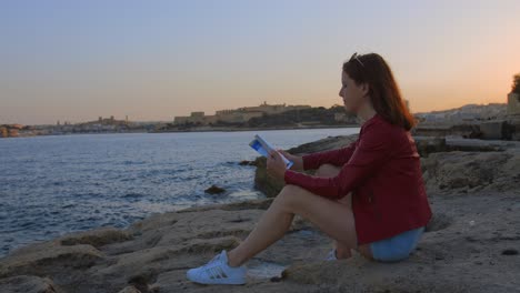 Eine-Junge-Frau-Sitzt-Mit-Einem-Buch-In-Der-Hand-Am-Strand-Am-Meer