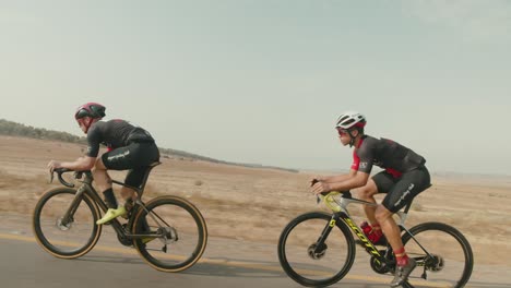 Toma-De-Seguimiento-En-Cámara-Lenta-De-Dos-Ciclistas-De-Carretera-Profesionales-Montando-Rápido-En-La-Carretera-Vacía-Del-Desierto