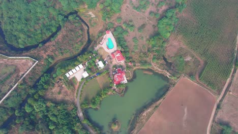 Toma-Aérea-De-Un-Dron-Súper-Alto-En-Vadodara,-India,-Sobre-Una-Casa-Con-Una-Piscina-Rodeada-Por-Un-Bosque-Verde-Y-Un-Lago,-Los-Campos-De-Cultivo-Parecen-Pequeños,-Como-Un-Mapa-Digital