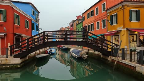 Pareja-Cruza-Un-Pequeño-Puente-De-Madera-Sobre-El-Canal-De-La-Isla-De-Burano-En-Italia-Con-Casas-Coloridas-En-El-Fondo