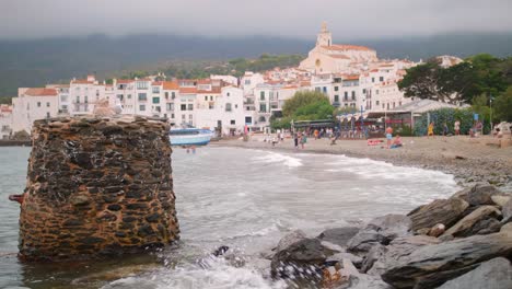 Turistas-En-La-Playa-De-Cadaqués,-España-Con-La-Iglesia-Católica-De-Santa-María-De-Cadaqués-En-Segundo-Plano.