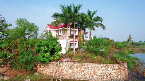 Aerial-drone-approach-to-white-vacation-house-and-boat-on-the-lake,-rising-towards-the-green-palm-trees-and-vegetation,-red-roof,-looking-up-at-the-resort-in-Vadodara,-India