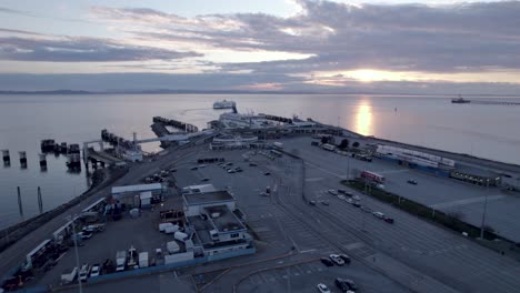 Drone-fly-above-parking-lot-at-bc-ferries-terminal-with-scenic-coastline-view-and-big-cargo-boat-cruising-the-calm-water-at-distance