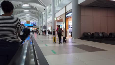 Woman-on-moving-sidewalk-inside-Istanbul-Airport-points-to-direct-husband-with-luggage-to-nearby-coffee-shop