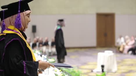 Happy-young-graduate-collecting-certificate-from-female-dean-with-friend-in-background