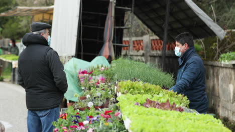 Hombre-Con-Chaqueta-Y-Mascarilla-Está-Vendiendo-Verduras-En-El-Mercado-Mientras-Habla-Con-Un-Cliente-Durante-La-Pandemia-En-Leiria,-Portugal