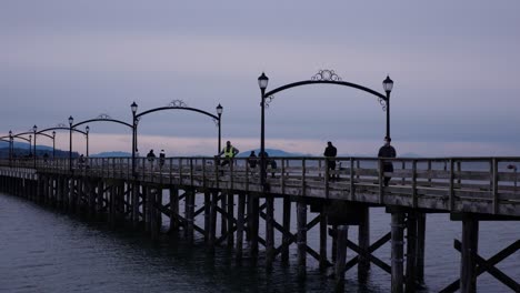 Vista-Estática-De-La-Gente-Caminando-En-Un-Largo-Muelle-Que-Conduce-A-Un-Rompeolas-En-El-Océano-Durante-Las-Nubes-De-La-Noche-Al-Atardecer