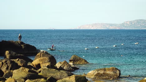 Fisherman-in-traditional-basket-boat-speaks-to-man-on-shore,-Vinh-Hy-Bay-Vietnam