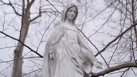 Estatua-De-Piedra-De-Mármol-Blanco-De-La-Madre-María-En-Un-Día-Sombrío-Con-Ramas-De-árboles-Ondeando-En-El-Fondo